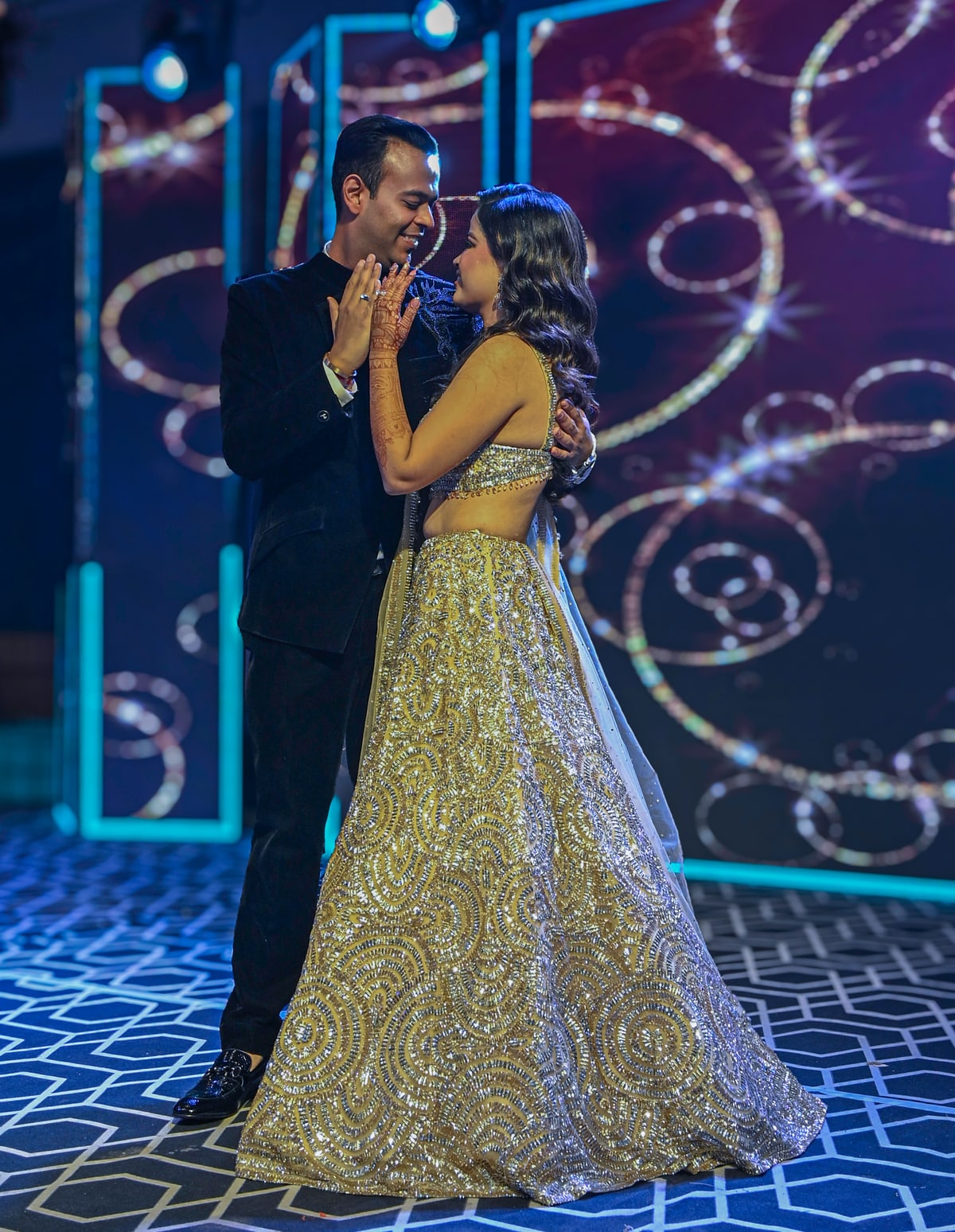 A bride in a gold lehenga and a groom in a black suit dance close on a glittering stage.
