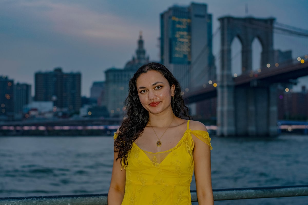 A woman in a yellow sundress smiles by the East River at dusk, the Brooklyn Bridge behind her.