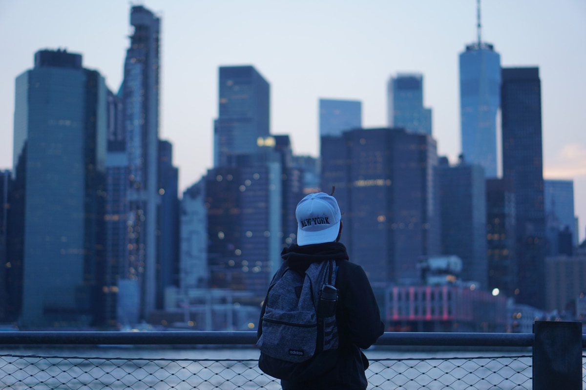 A person in a New York cap and backpack looks out at the Lower Manhattan skyline at dusk.