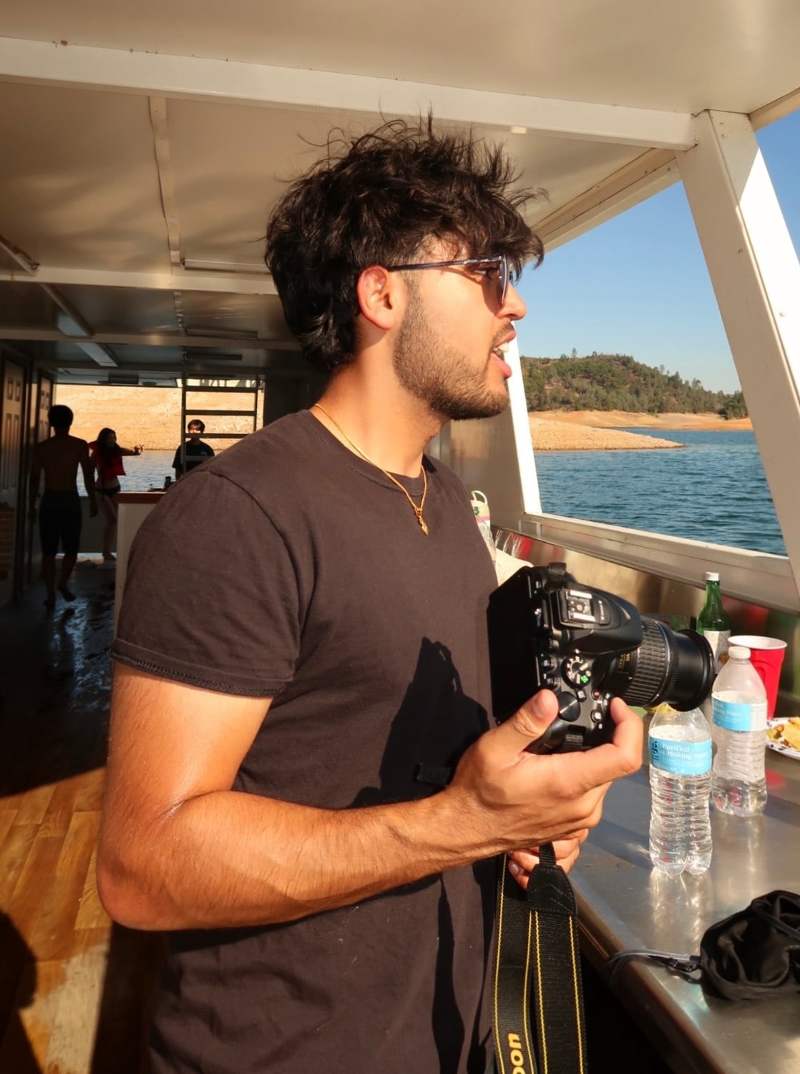 Smayan Gupta holding a Nikon camera on a boat deck on a sunny lake.
