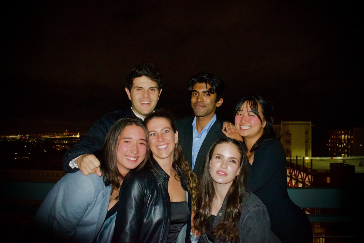 Six friends squeeze into the frame on a rooftop with the city glowing behind them.