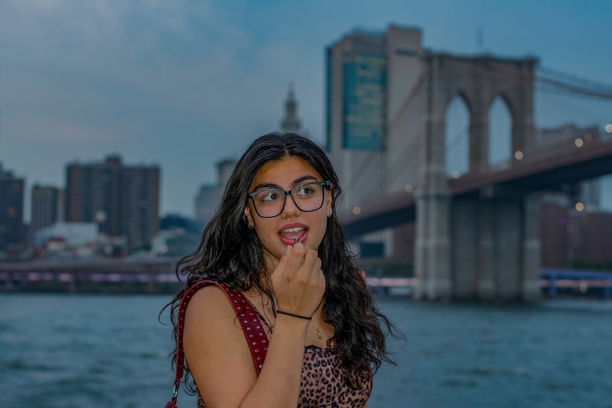 A woman in glasses with curly hair poses by the East River with the Brooklyn Bridge behind her.