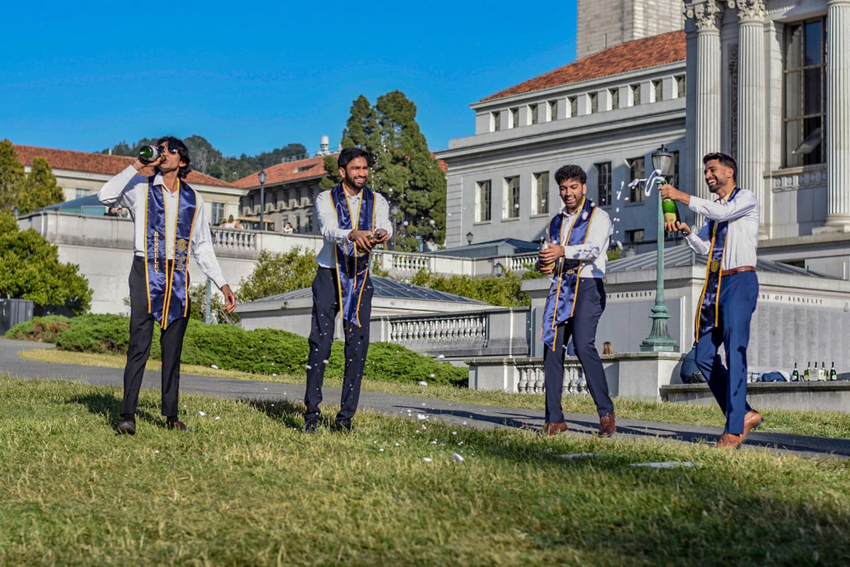 Graduates spray champagne on a sunny lawn in front of a domed campus building.