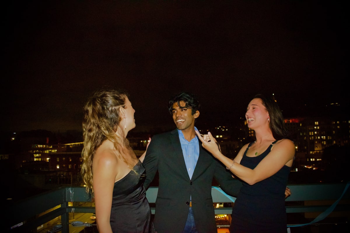 Two women in black dresses laugh with a friend in a suit at the rooftop railing.
