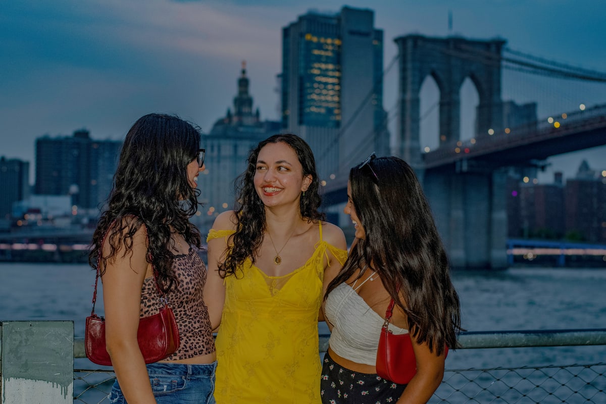 Three friends laugh together on the Brooklyn waterfront at dusk.