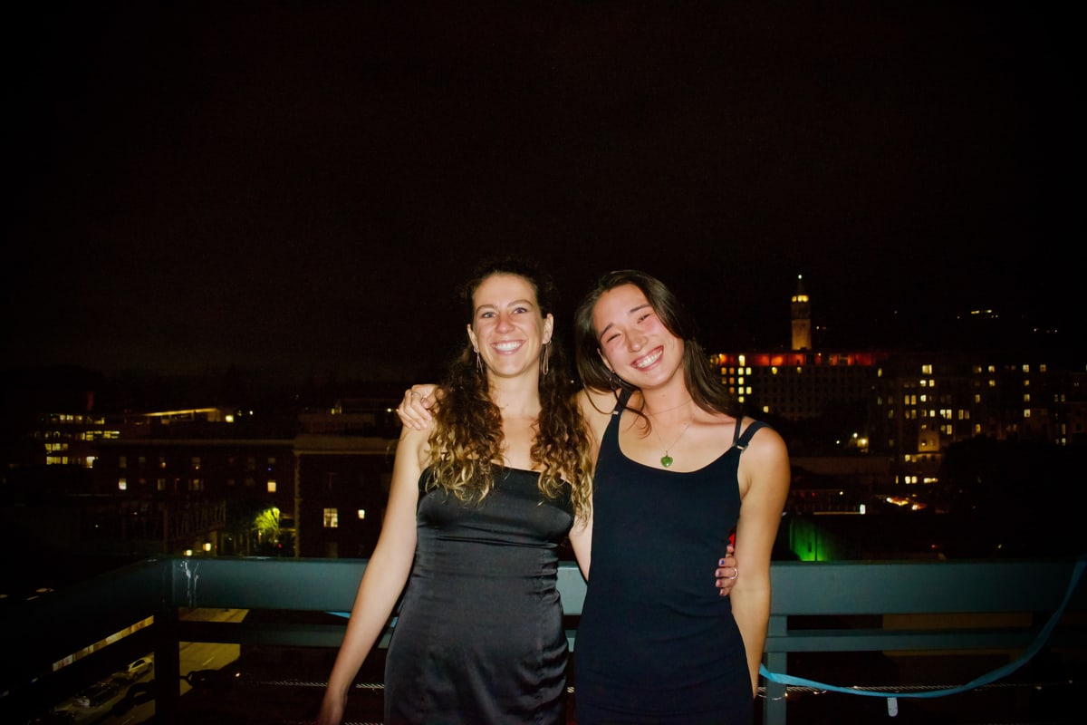 Two friends in black dresses pose arm in arm with city lights and a clock tower behind them.