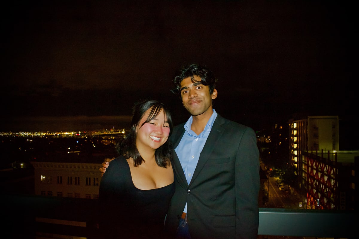A smiling couple stands at the rooftop railing above the city at night.