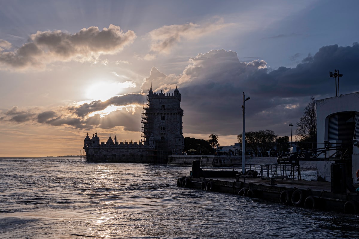 Belém Tower in silhouette as the sun breaks through clouds over the river.