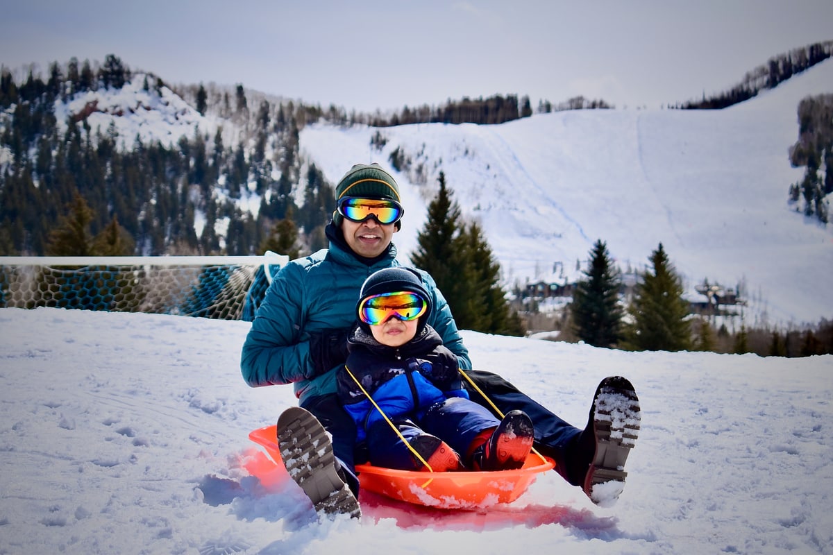 A father and young child in ski goggles ride a sled together.