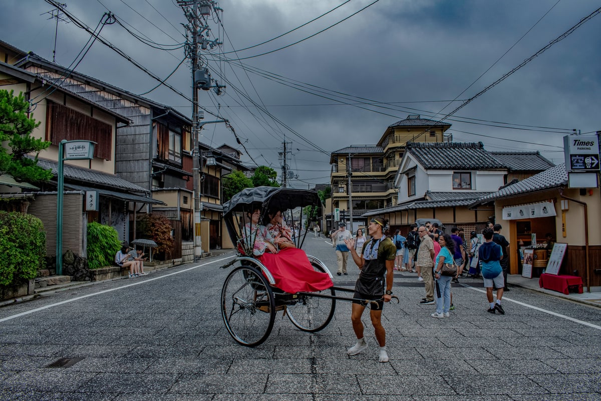 A rickshaw driver pulls passengers down a quiet Kyoto street under grey skies.
