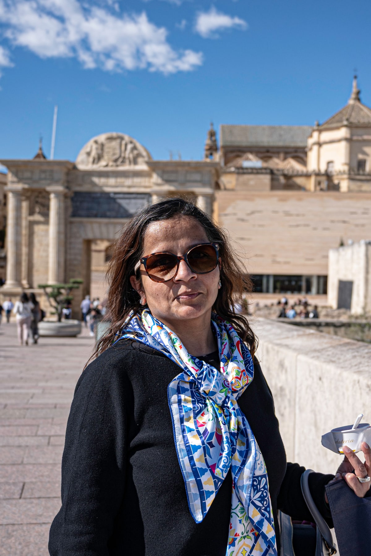 A woman in sunglasses and a patterned scarf smiles in front of historic stone buildings.
