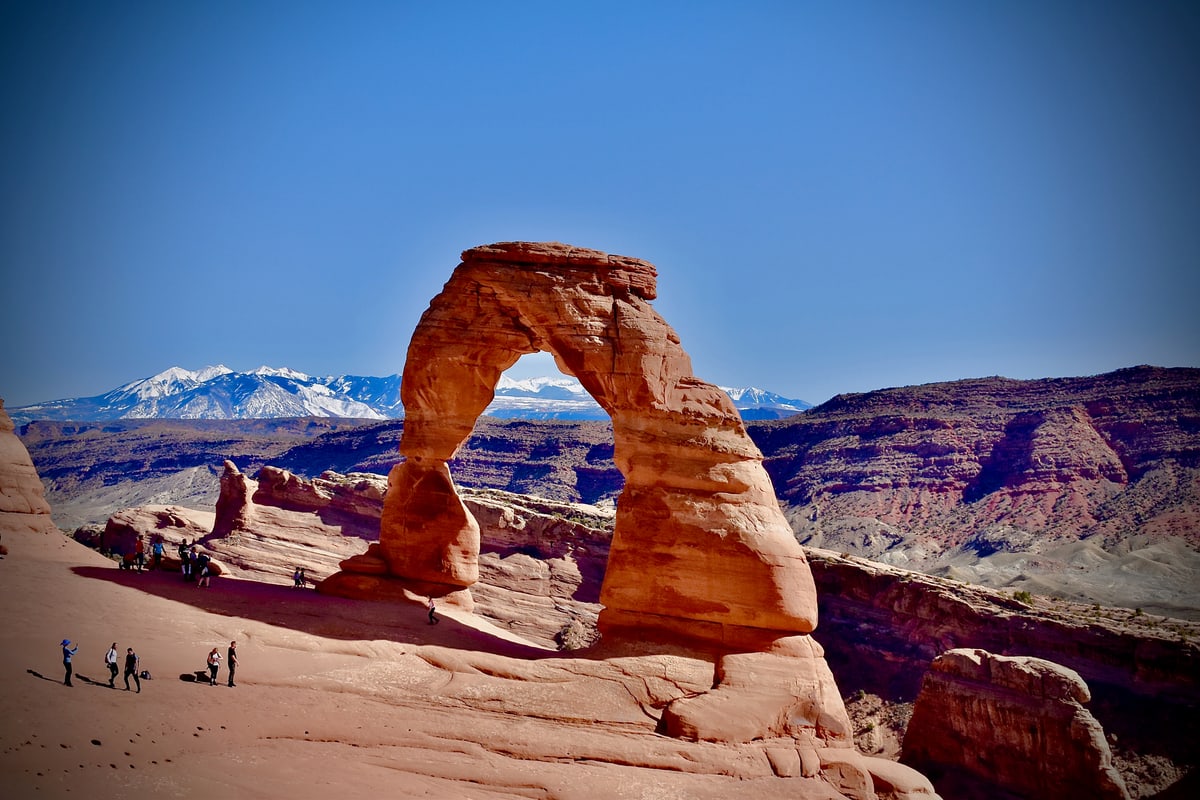 Delicate Arch stands against a blue sky with snowy mountains in the distance.