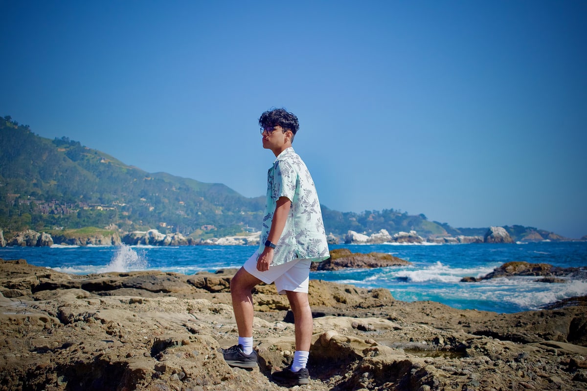 A young man in a floral shirt stands on coastal rocks as waves break behind him.