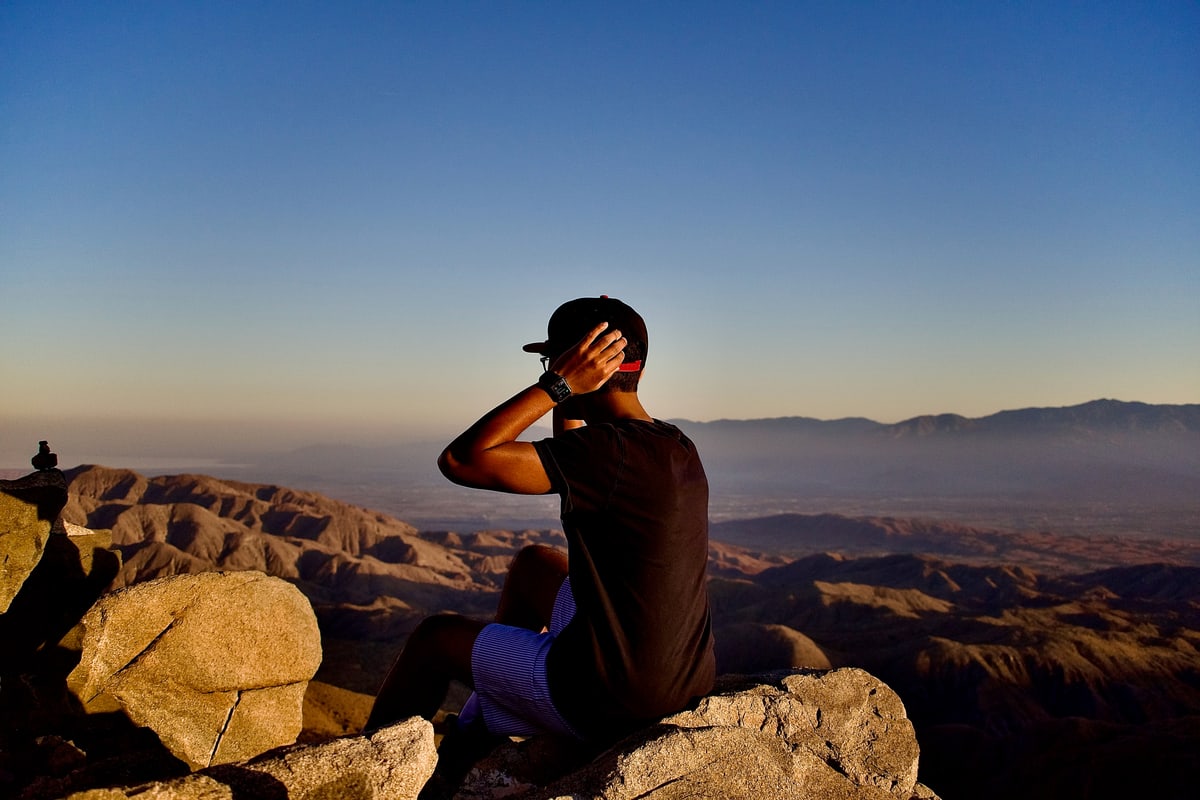 A man sits on a boulder looking out over desert mountains at dusk.