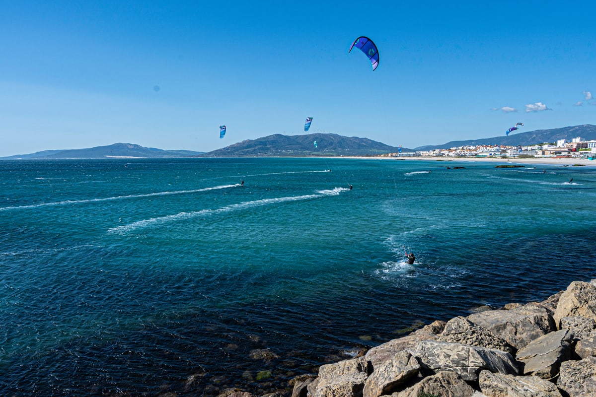 Kitesurfers skim across deep blue water off Tarifa with mountains beyond.