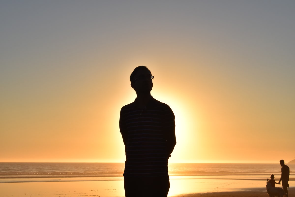 A man's silhouette stands against the setting sun on a beach.