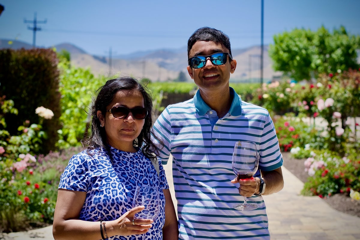 A smiling couple in sunglasses stands in a rose garden, one holding a glass of wine.