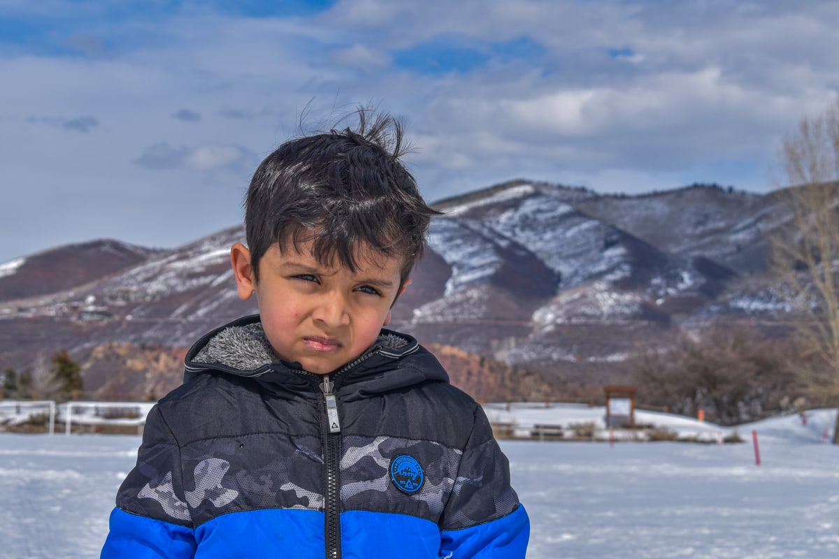 A young boy in a puffy jacket squints in the snow with mountains behind him.
